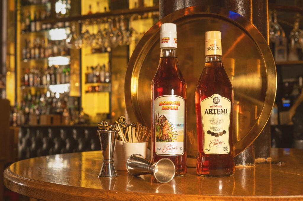 Two glistening golden bottles atop a table in a bar. The first is Ron Miel Indias and the second is Artemi Ron Miel Honey Rum.